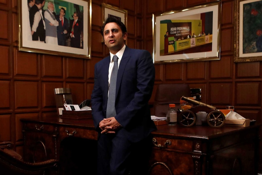 A man in a suit leans against the desk in an office with wood-panelled walls