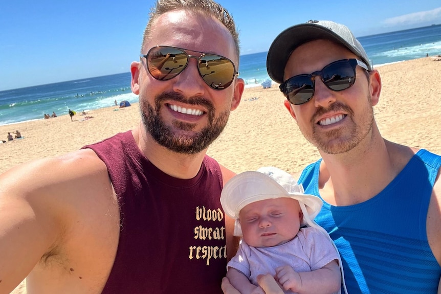 Two men in singlets and sunglasses pose for a photo with a little baby on the beach