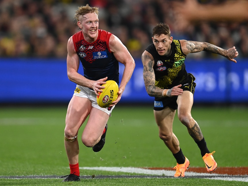 Melbourne AFL player Clayton Oliver smiles as he runs with ball in hands, looking upfield as a defender tries to get to him.