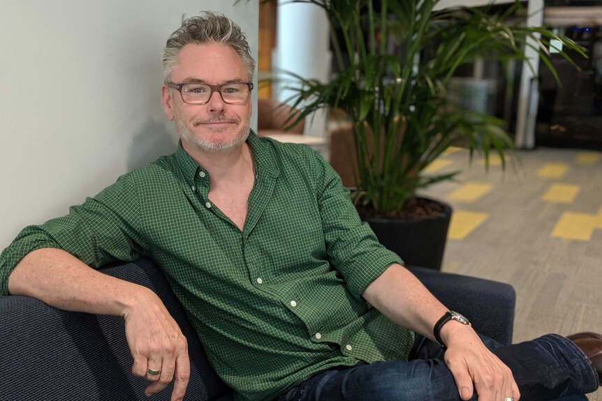 A man with grey hair and glasses and green shirt leans back on a couch and looks seriously at the camera