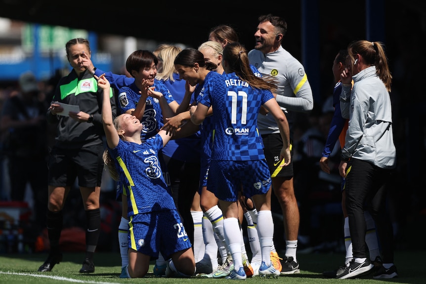 A group of female soccer players in blue celebrate in a huddle during a game with smiles on their faces