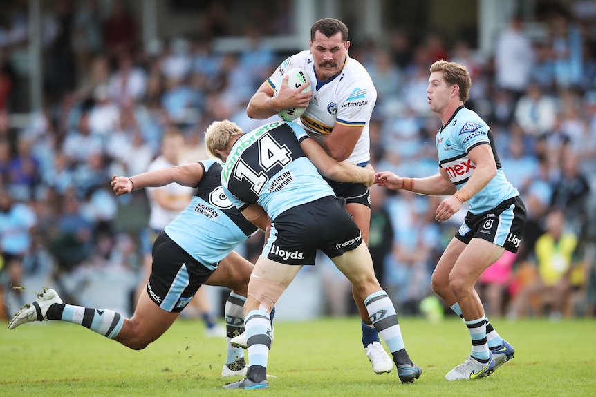 Un jugador de la NRL sostiene el balón y se para en un placaje mientras tres jugadores de la oposición intentan derribarlo.