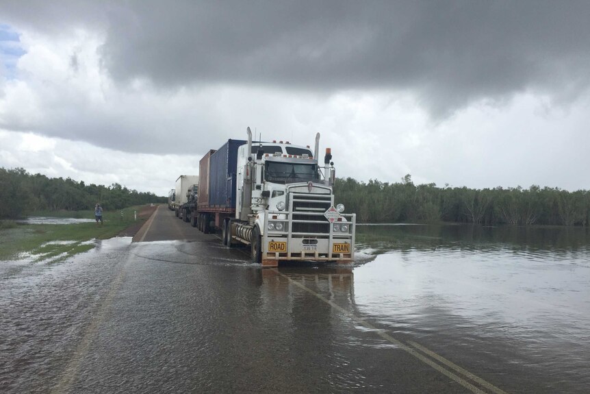 A road train drives through floodwater on the Great Northern Highway at Roebuck Plains under grey skies.