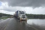 A road train drives through floodwater on the Great Northern Highway at Roebuck Plains under grey skies.