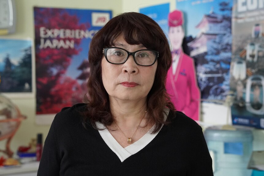 A woman stands in an office with travel posters behind.