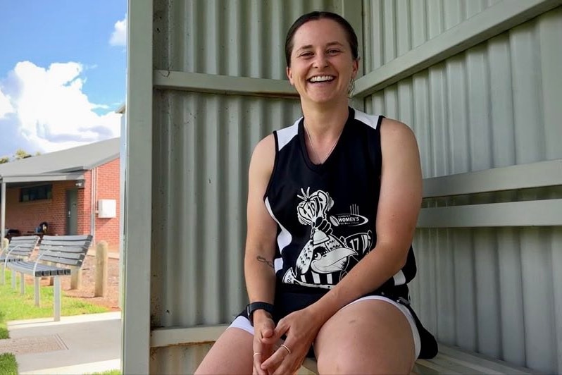 woman in footy outfit sits in dug out on side of oval, smiling at camera