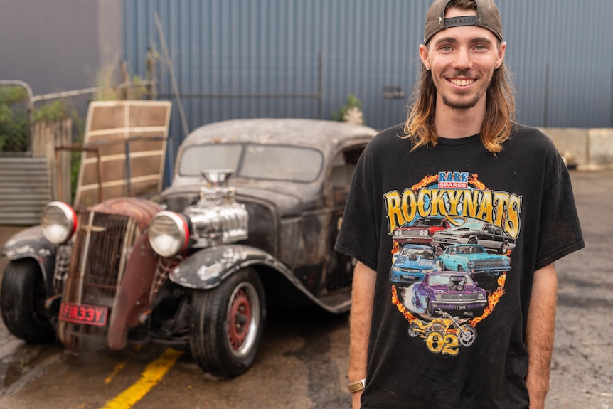A young man with long hair and hat backwards with a black car T-shirt standing in front of a hot rod.