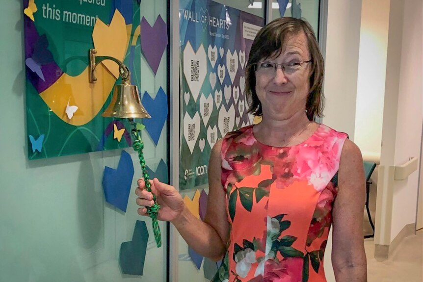 A woman in a colourful dress smiles as she rings a brass bell mounted on the wall