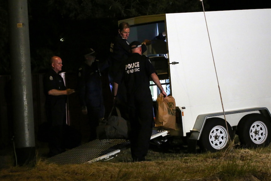 A close-up shot of three police forensic officers standing at the rear of a white box trailer at night.