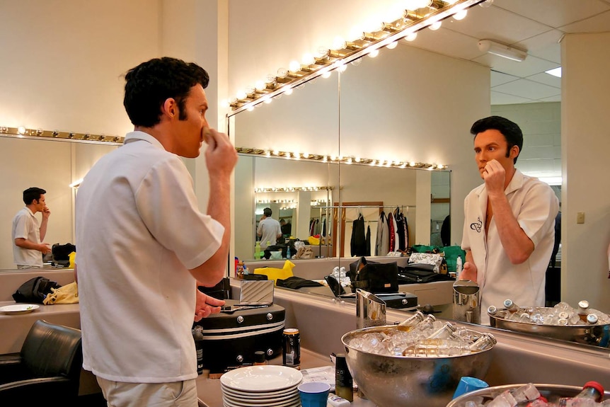 Elvis tribute artist Brody Finlay applies face make up in a dressing room with rows of illuminated lights.