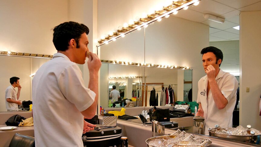 Elvis tribute artist Brody Finlay applies face make up in a dressing room with rows of illuminated lights.