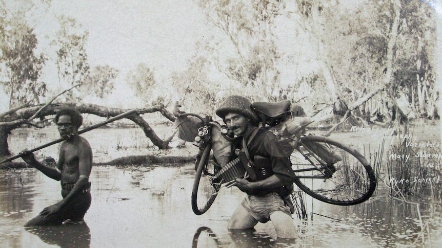 A black and white photo of a non-Indigenous man walking through a swamp with his bike near an Indigenous man with a spear.