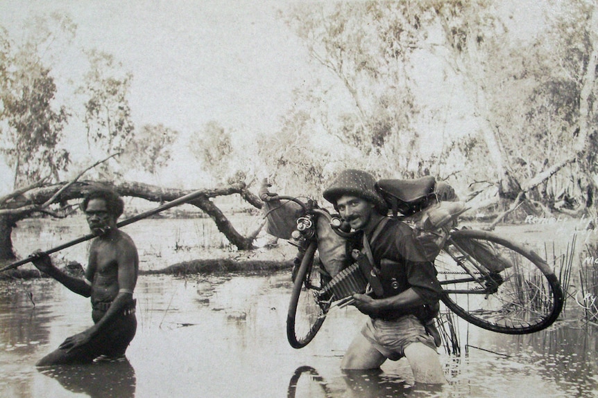 A black and white photo of a non-Indigenous man walking through a swamp with his bike near an Indigenous man with a spear.