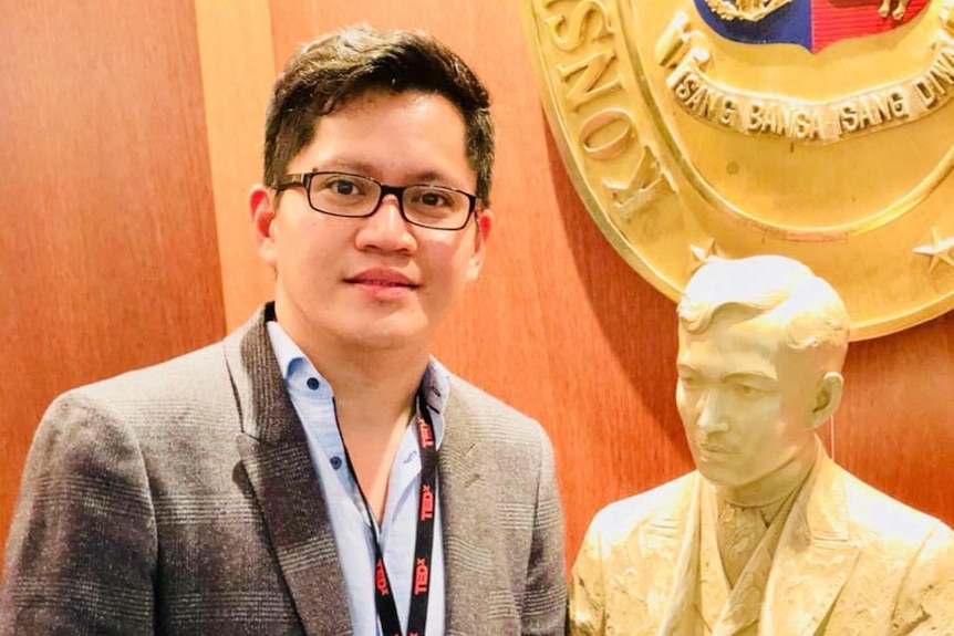 Rado Gatchalian wears a suit posing next to a statue of a man's head. 