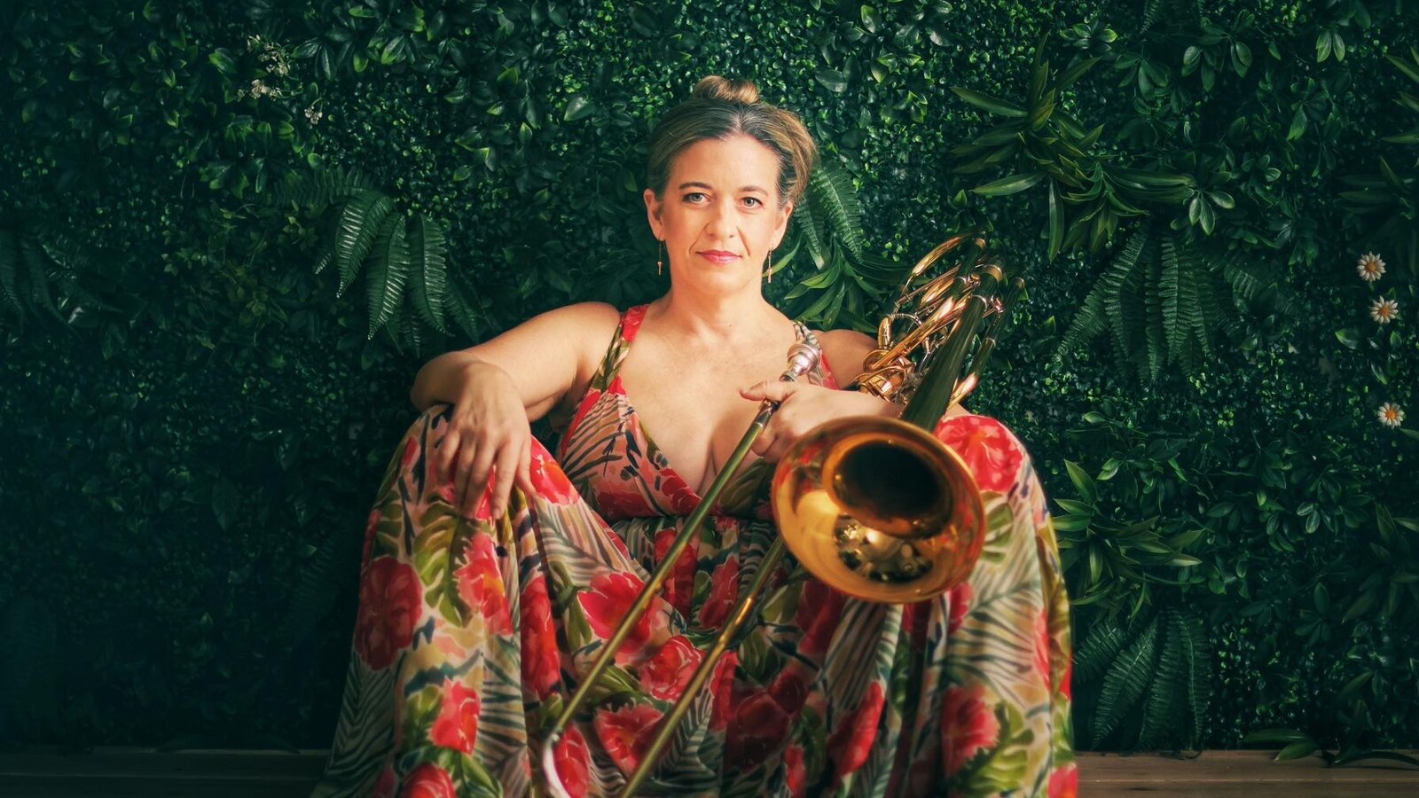 Trombonist Jennifer Wharton sits in a floral dress in front of a green wall covered in ferns; she's holding her bass trombone