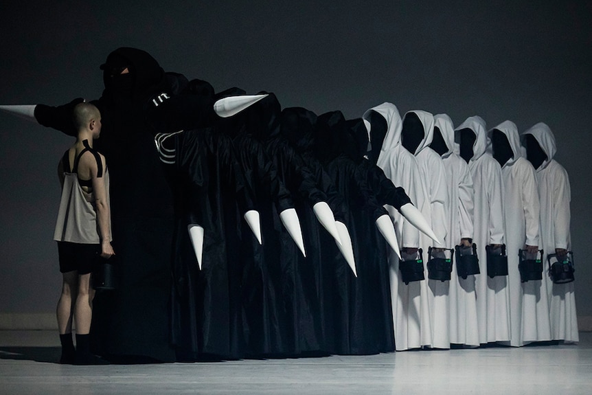 Single dancer stands facing line of 14 dancers one in front of the other, half in black robes and half in white, faces covered.
