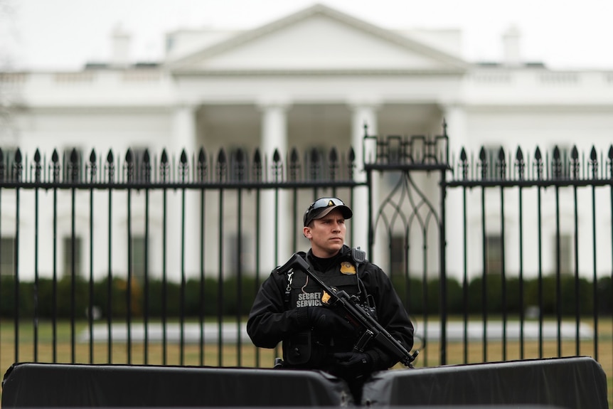 A male secret service officer in uniform holds a gun as he stands with his back to a fence, in front of the White House