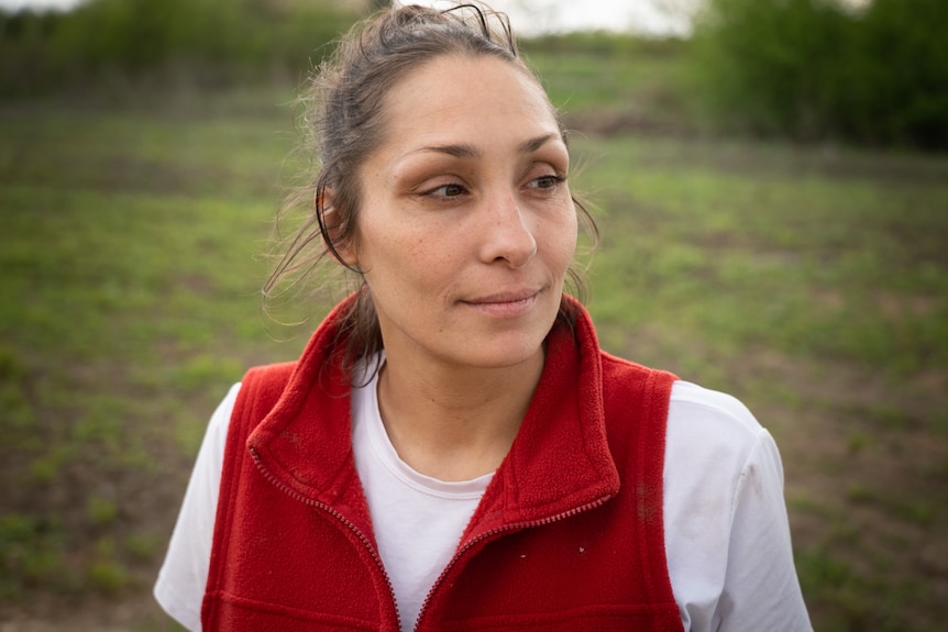 A woman with her hair tied in a pony tail and wearing a red vest and shirt looks away from the camera 
