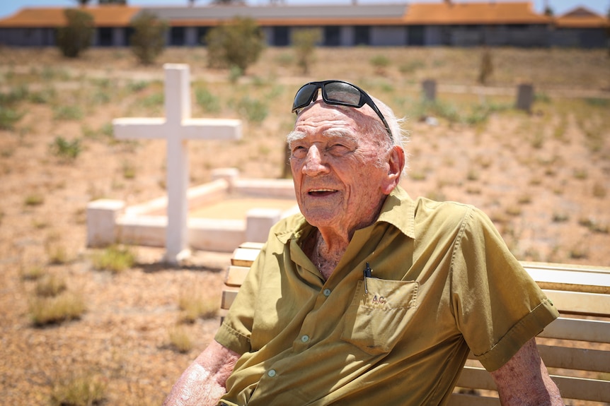 A smiling elderly man sits on a bench in a cemetery, with a cross headstone behind him.