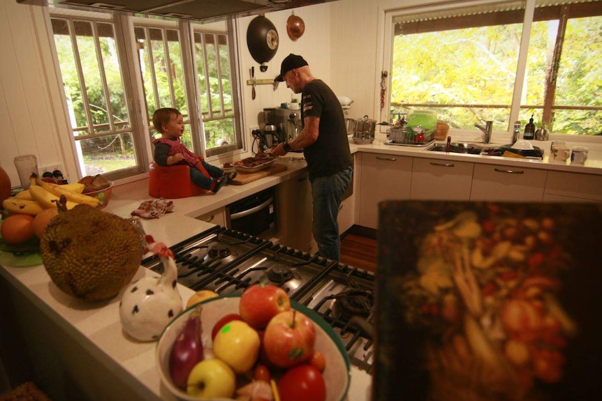 A man in a kitchen with his back turned to the camera as he prepares food with a toddler sitting in a chair on the bench