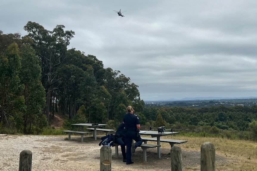 A helicopter flying in the air with police standing near a picnic table with a drone on it.