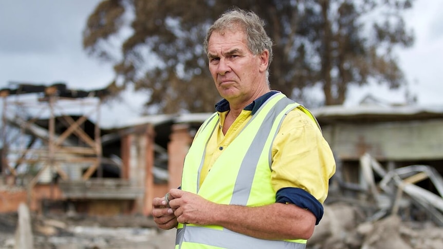 A man in jeans, a yellow tradesman's top and high vis vest looks solemn. A burnt out building can be seen in the background.