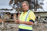A man in jeans, a yellow tradesman's top and high vis vest looks solemn. A burnt out building can be seen in the background.