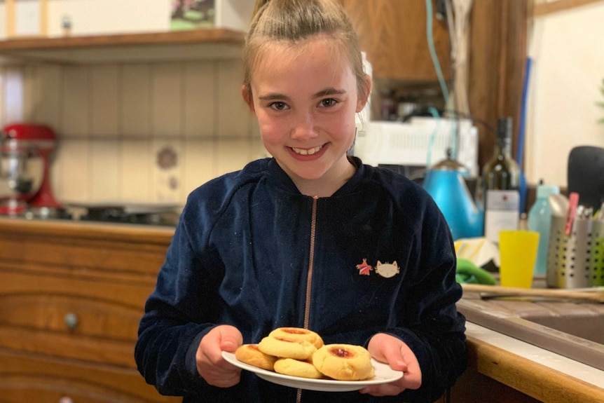 A girl standing in a kitchen holding a plate of jam-drop biscuits and smiling.