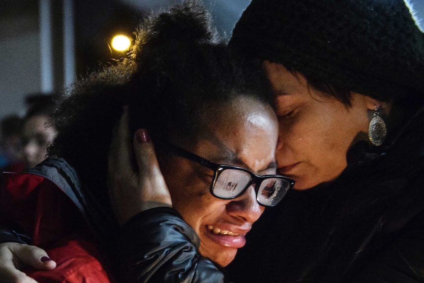 A woman grimaces in distress, weeping as another woman holds her close, her hand cradling her head.