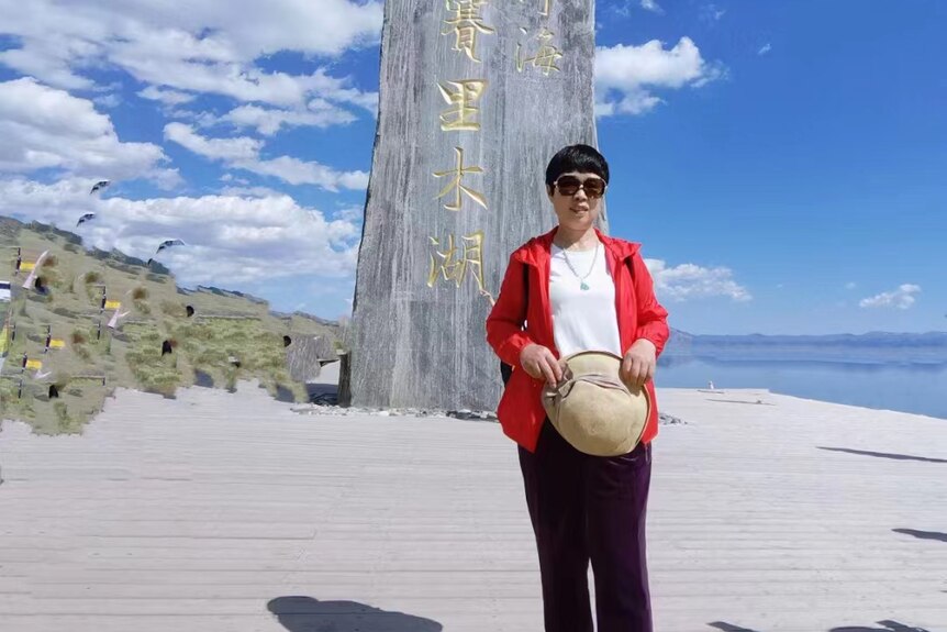 A woman wearing black pants and a red jacket stands on a white sand beach next to the ocean