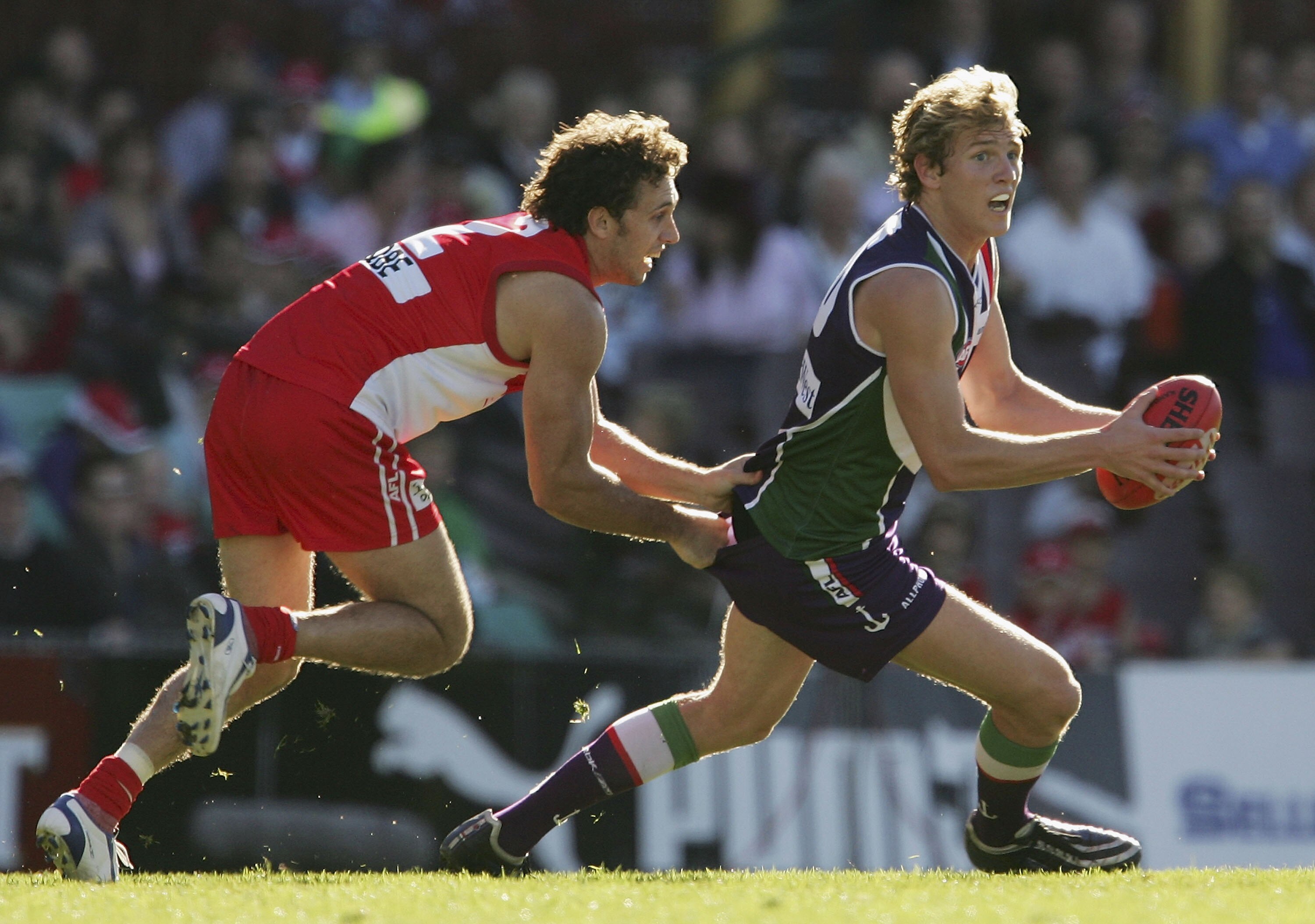 A Fremantle player holds the ball in front of him as he looks downfield while a Sydney player grabs onto his shirt.