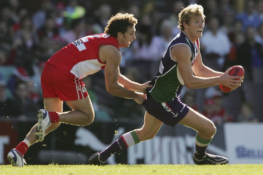 A Fremantle player holds the ball in front of him as he looks downfield while a Sydney player grabs onto his shirt.
