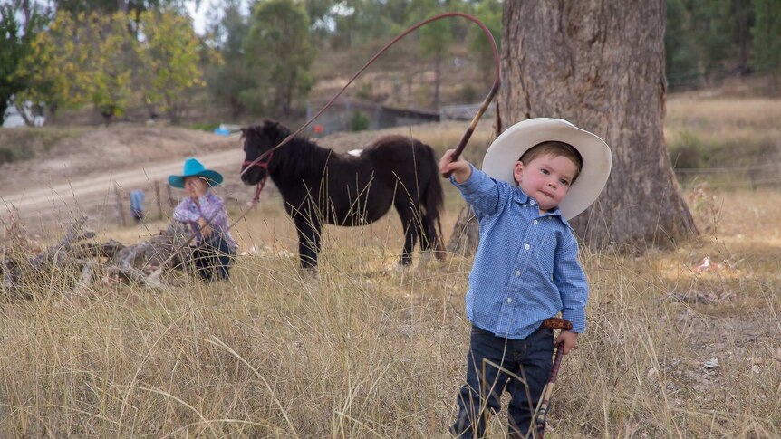 A toddler boy in a blue check shirt and cowboy hat cracking a whip in front a tree and horse and girl in the background