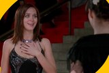 Emily holds her hand up to her chest and looks excited as she stands on steps wearing a fancy gown with Andie facing her.