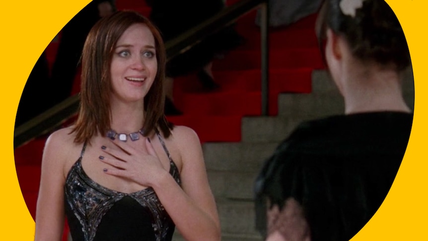 Emily holds her hand up to her chest and looks excited as she stands on steps wearing a fancy gown with Andie facing her.