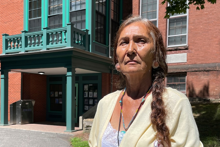 A woman with braided hair wearing native american jewellery stands looking upp at the sun in front of a brick building.