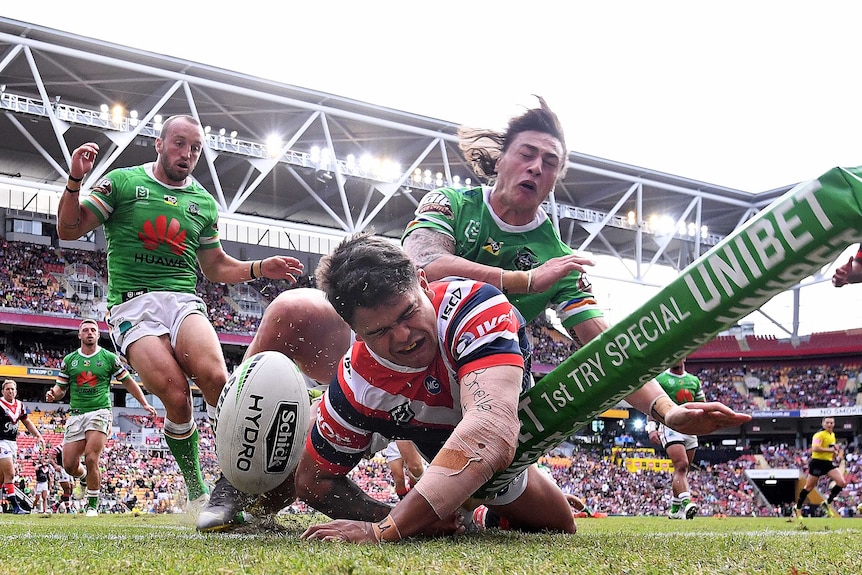 An NRL player grimaces as he scores in the corner as an opposition player lands on him.