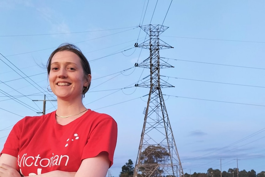 A woman, wearing a red T-shirt with her arms crossed, standing in front of a large electricity pole.