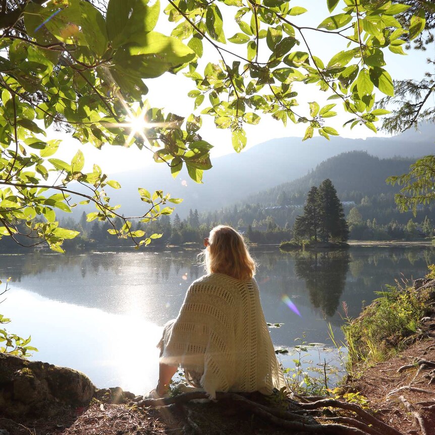 Blonde woman sits gazing over peaceful lake as the sun rises with mountains in background