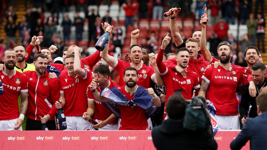 A group of Wrexham footballers in red shirts stand together cheering with arms raised in triumph. 