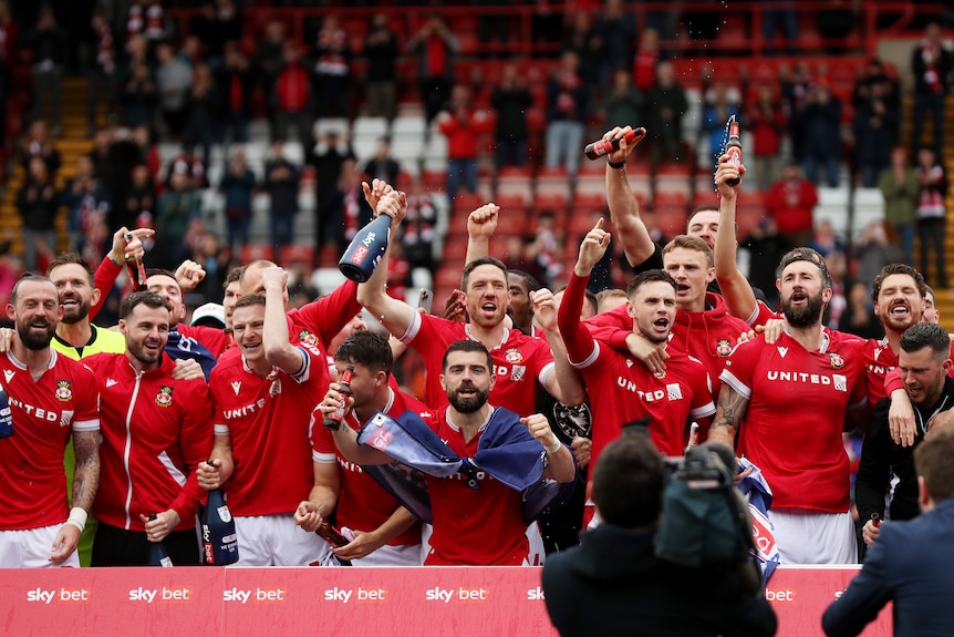 A group of Wrexham footballers in red shirts stand together cheering with arms raised in triumph. 