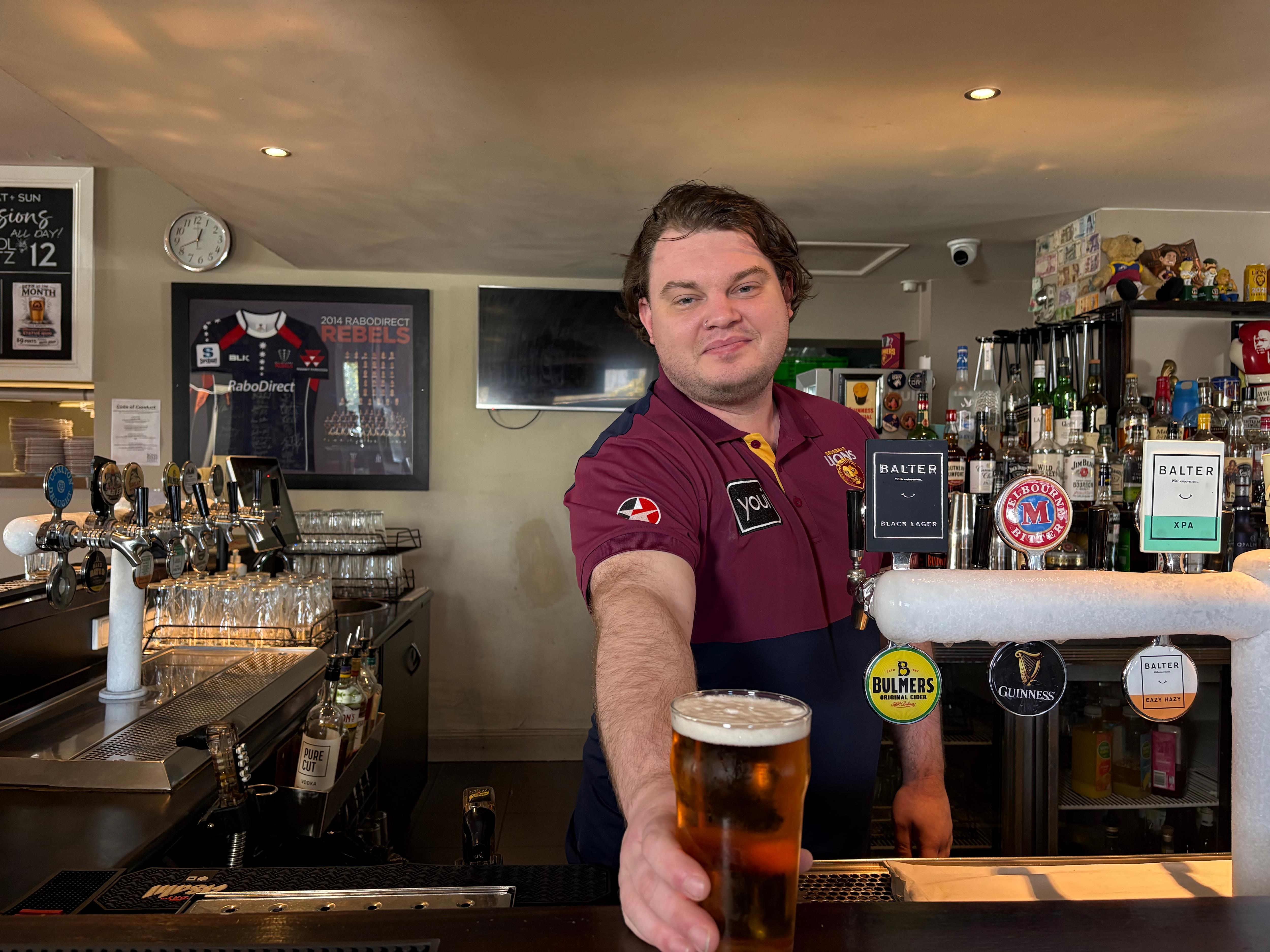 A dark-haired man wearing a Brisbane jersey passing a freshly poured beer over the bar.