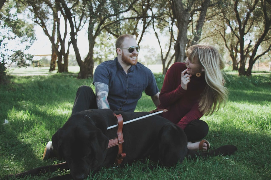 A wide shot of Arron and Morgan sitting outside on a picnic rug with a seeing eye dog in the foreground.