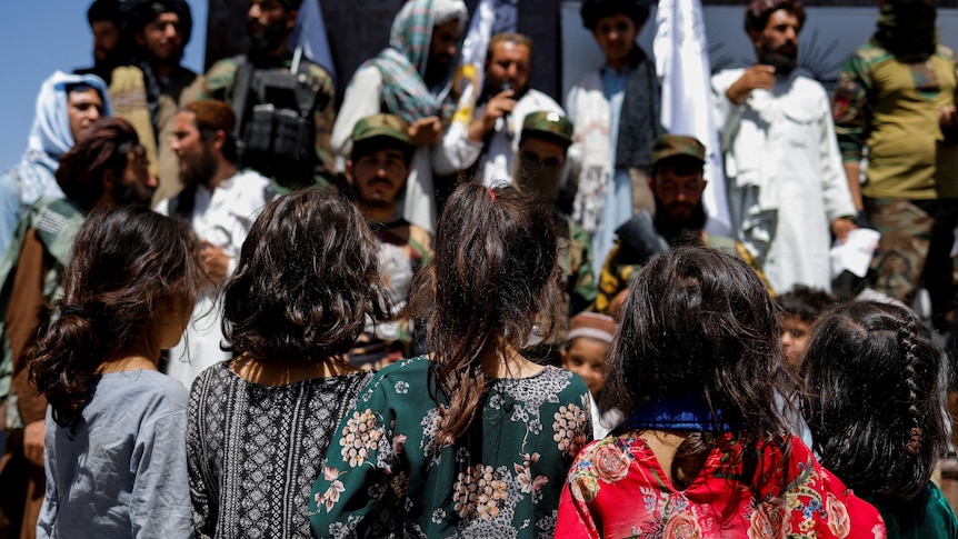 The backs of four young Afghan girls facing men perched above them who are Taliban supporters.