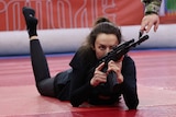 A woman lays on the ground holding a gun and ponting it straight ahead. 