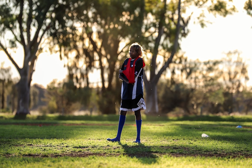Young Aboriginal woman stands wearing football clothing and socks on sun drenched football oval