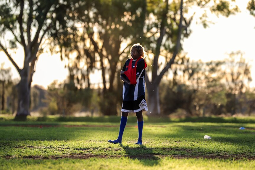 Young Aboriginal woman stands wearing football clothing and socks on sun drenched football oval