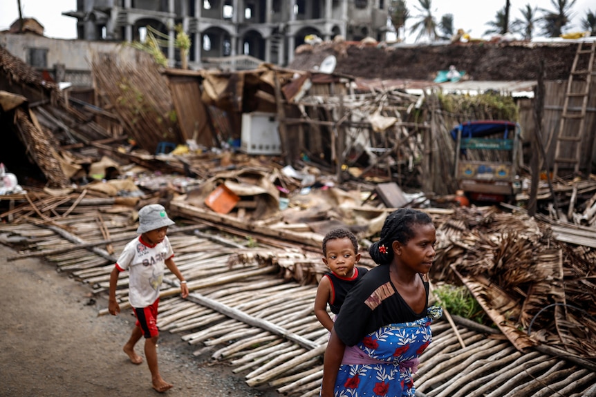 A woman carries a young child, and another follows, as they walk past destroyed houses