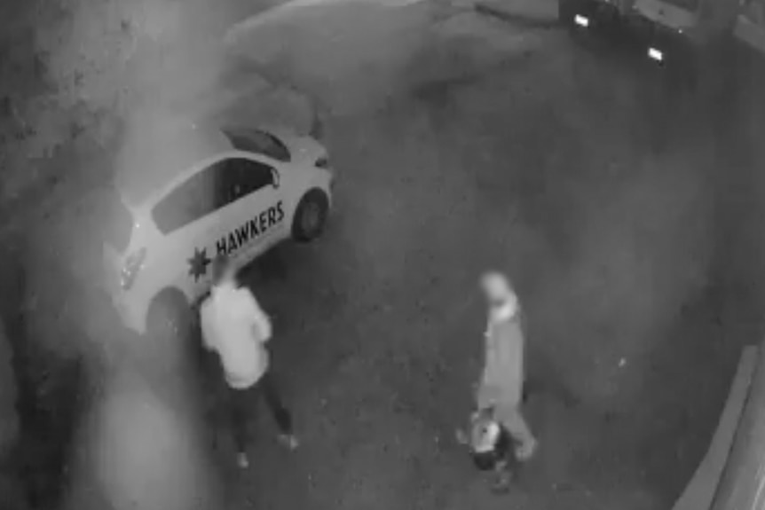 Black and white CCTV footage of two men carrying beer kegs with a car parked nearby