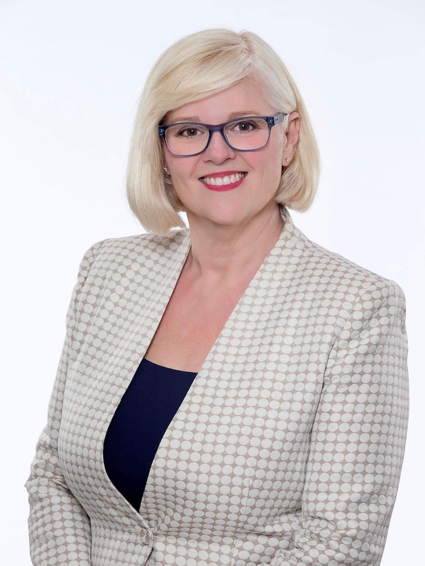 A blonde woman wearing purple-rimmed glasses smiles for a portrait wearing a checked blazer.
